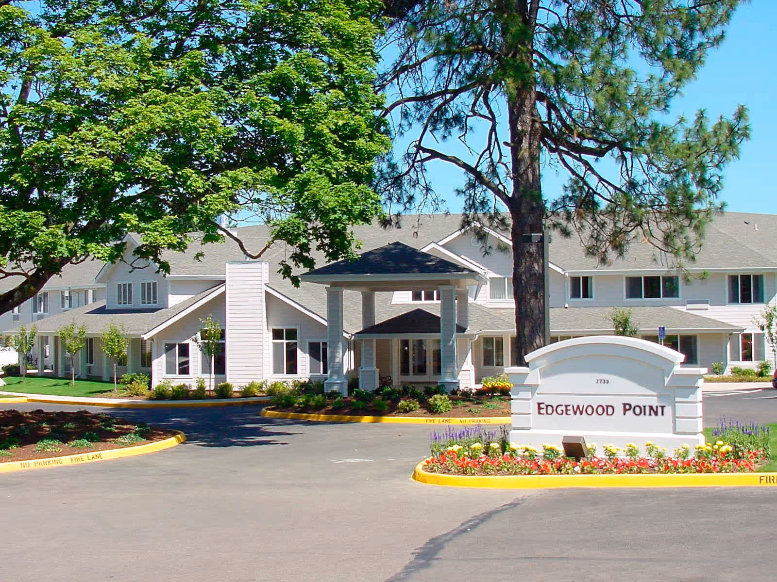 Exterior view of Edgewood Point Senior Living facility showing a large white building with multiple windows, a covered entrance, surrounded by trees and landscaped flower beds. A white sign with the name 'Edgewood Point' is prominently displayed in front of the building.