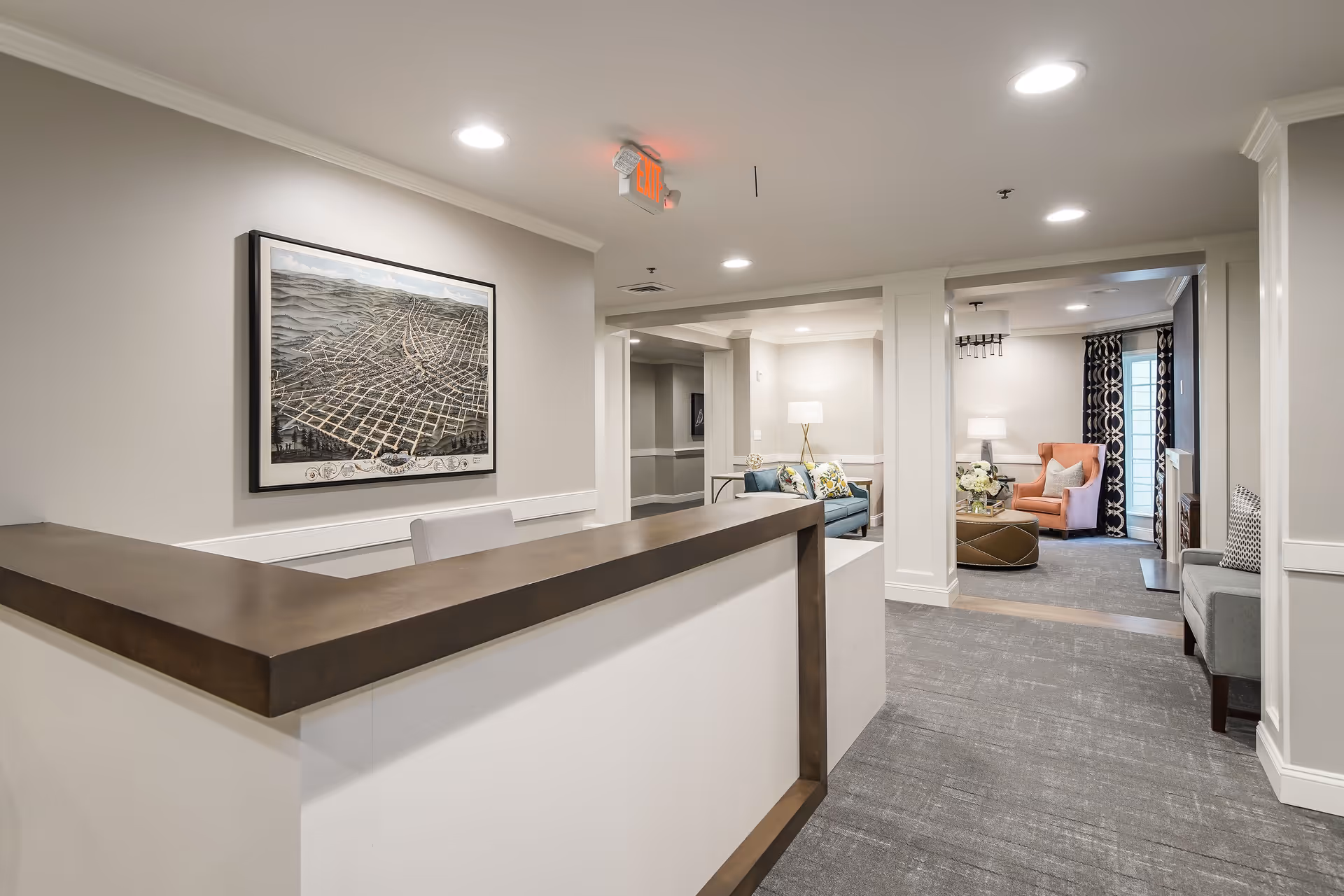 Interior view of a senior living facility reception area with a wooden reception desk in the foreground, a framed map on the wall, and a seating area with chairs, a sofa, lamps, and a coffee table in the background.