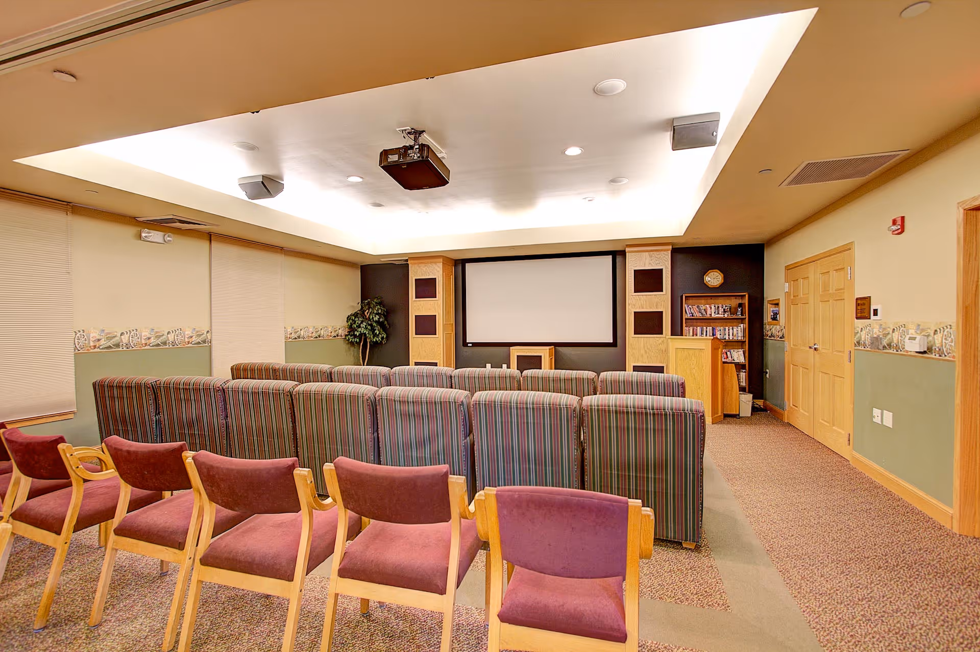 A small communal media/theater room with rows of upholstered chairs facing a large projection screen and a ceiling-mounted projector.