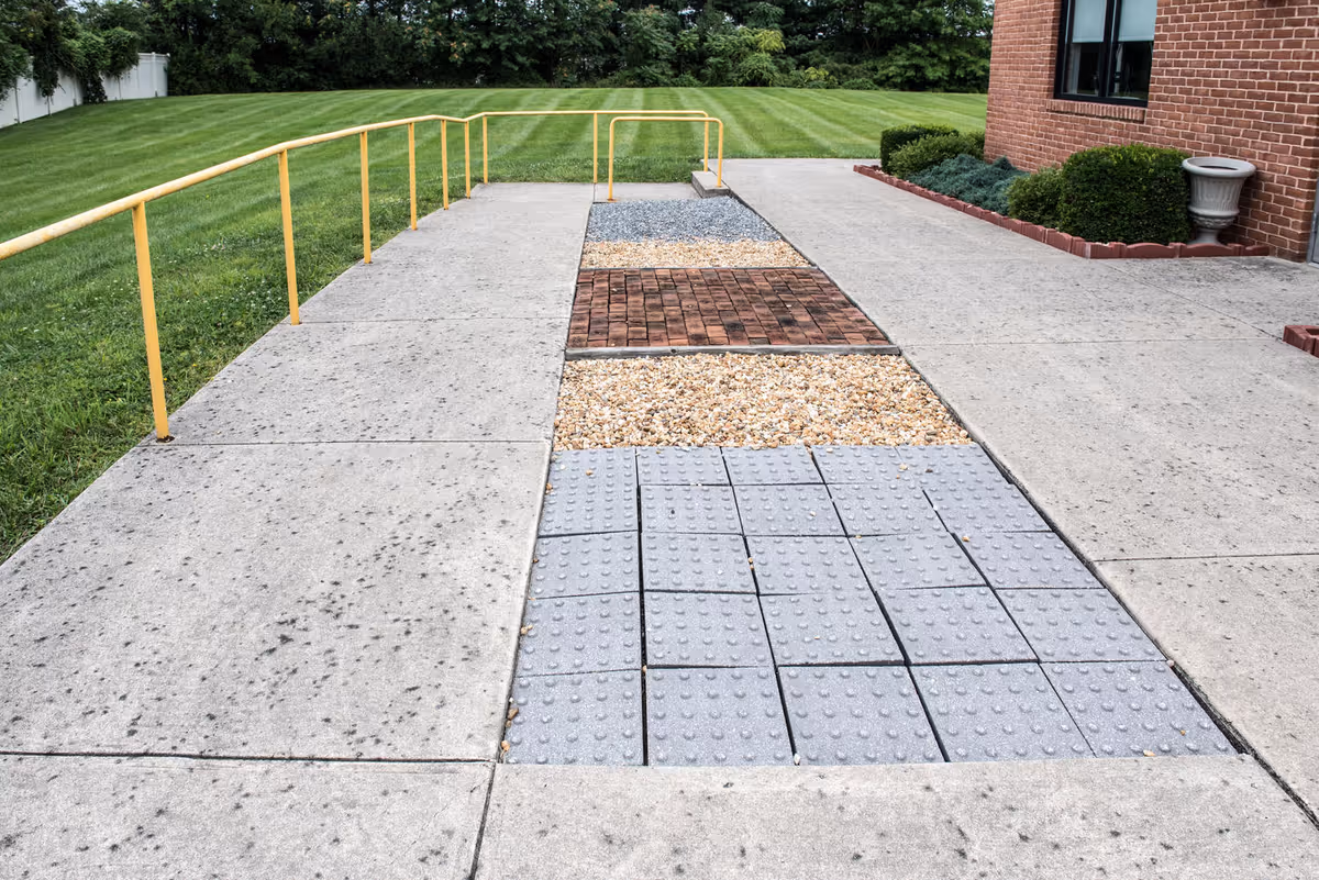 Outdoor concrete walkway with different textured sections including tactile paving, gravel, and bricks, bordered by a yellow handrail on the left side and a brick building with shrubs on the right side, with a well-maintained grassy area and trees in the background.