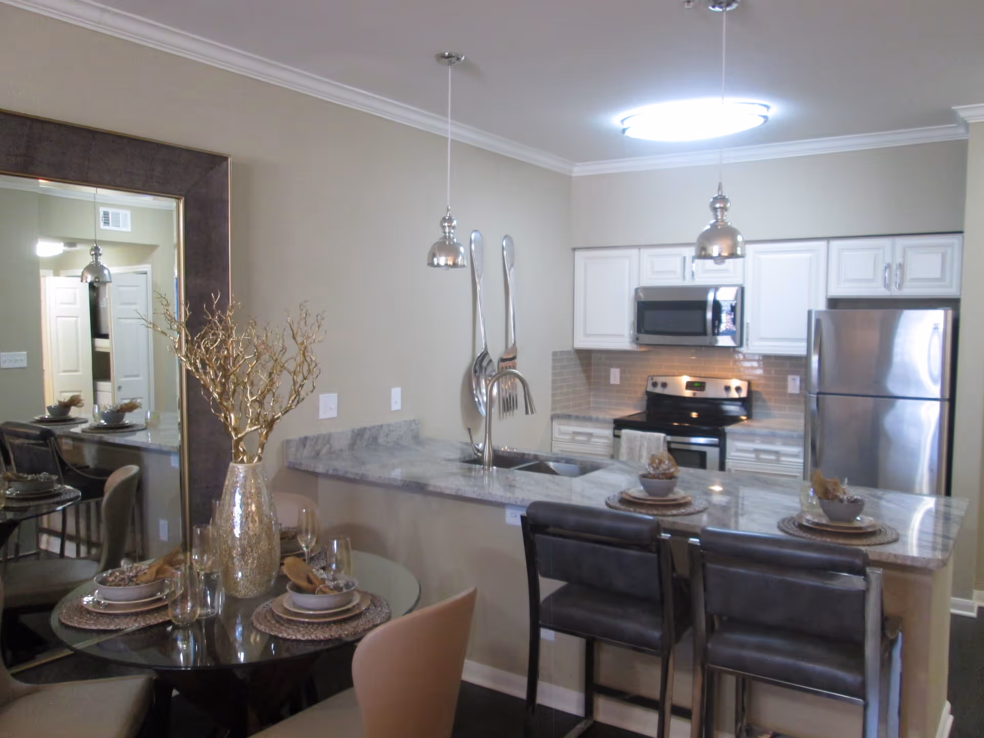 Interior view of a modern kitchen and dining area in an active adult living facility. The kitchen features white cabinets, stainless steel appliances including a refrigerator, stove, and microwave, and a marble countertop with a sink and two pendant lights hanging above. Adjacent to the kitchen is a round glass dining table set with four place settings and a decorative gold vase with branches. Two bar stools are positioned at the kitchen counter. A large mirror is mounted on the wall behind the dining table.