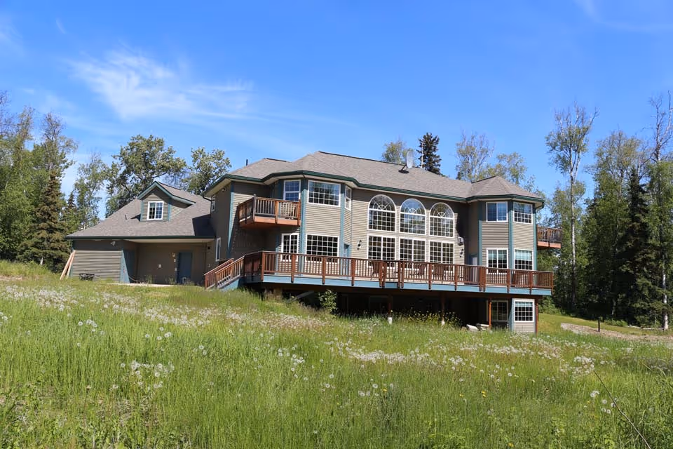 A large two-story house with multiple windows and balconies, surrounded by green grass and trees under a clear blue sky.