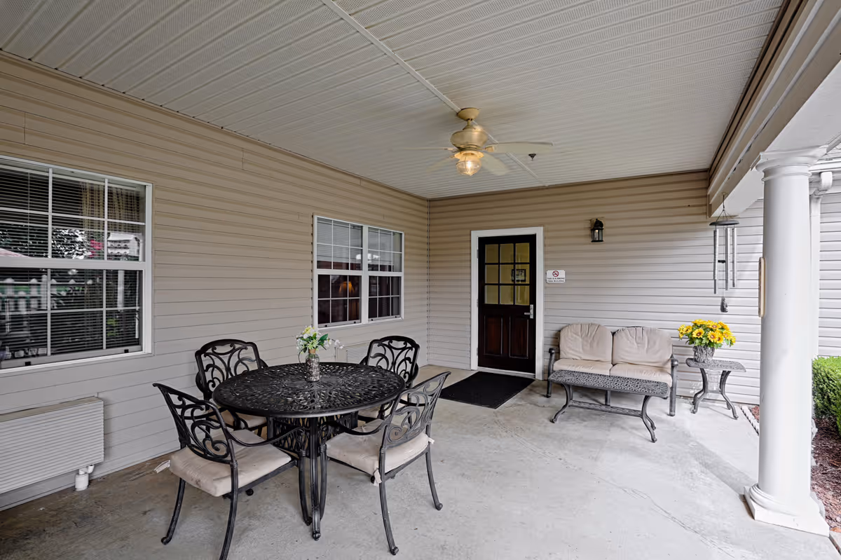 Covered outdoor patio area with a round black metal table and four matching chairs with beige cushions. A small vase with flowers is on the table. To the right, there is a cushioned bench with beige cushions and a small side table holding a vase of yellow flowers. The area has beige siding walls, two windows, a dark wooden door, a ceiling fan with light, and a white column supporting the roof.