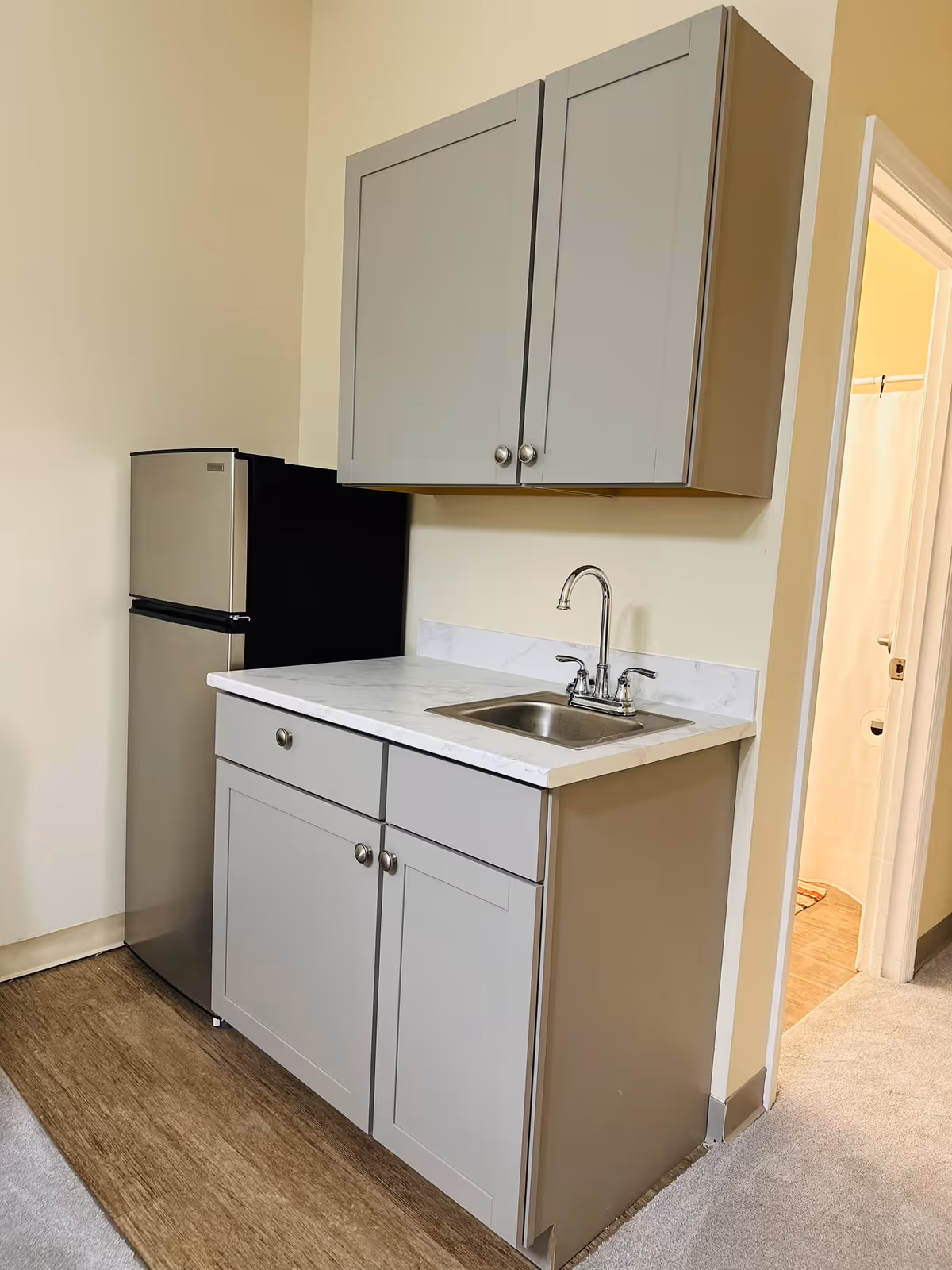 Small kitchenette area with a stainless steel mini refrigerator, gray cabinets, a white countertop, and a stainless steel sink with a faucet. To the right, there is an open doorway leading to a bathroom with a shower curtain visible.
