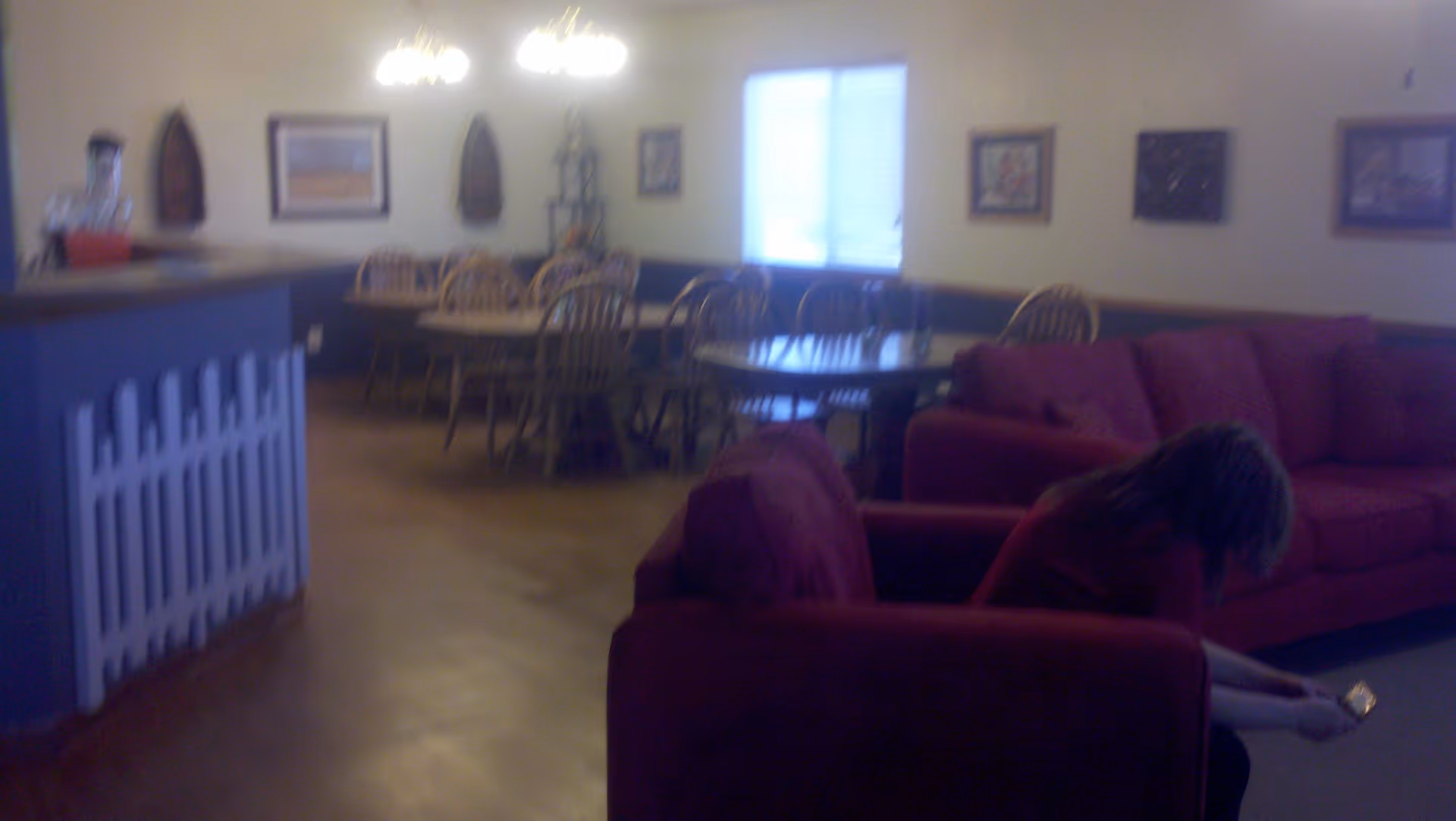 Interior of a senior living facility showing a common area with red couches in the foreground and wooden dining tables with chairs in the background. A person is seated on one of the couches looking down at a phone. The walls are decorated with framed pictures and there is a window with blinds letting in natural light.
