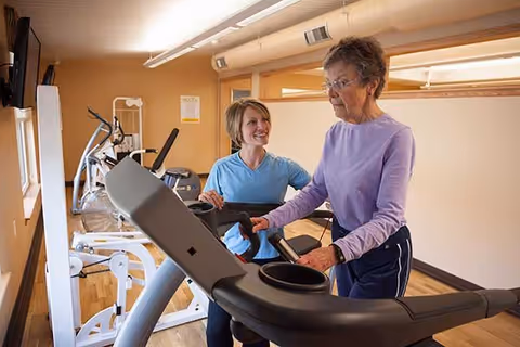 An elderly woman in a purple long-sleeve shirt and dark pants is walking on a treadmill while a younger woman in a blue shirt smiles and assists her in a fitness room with exercise equipment and wooden flooring.