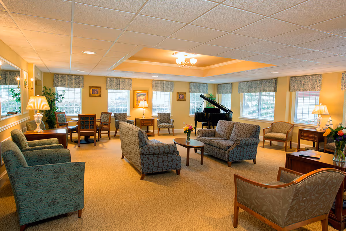 A spacious, well-lit living room with multiple upholstered chairs and sofas arranged around small wooden tables. There are several windows with patterned valances letting in natural light. A black grand piano is positioned in the corner near the windows. The room has beige carpeting, yellow walls, and several lamps providing warm lighting. Decorative plants and framed artwork adorn the walls.