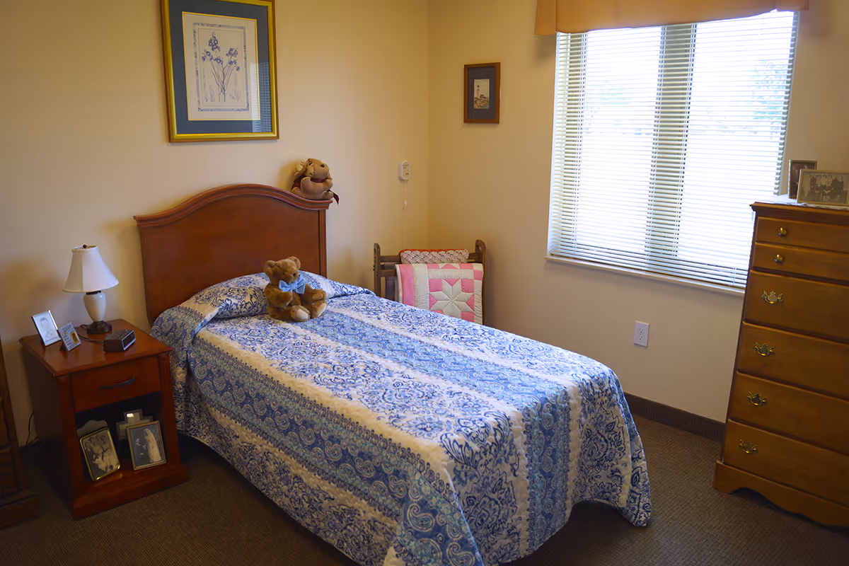 A cozy bedroom with a single bed covered in a blue and white patterned bedspread. A teddy bear is placed on the bed. There is a wooden nightstand with a lamp, framed photos, and a clock. A wooden dresser is positioned near a window with blinds, and a quilt rack with a pink and white quilt is in the corner. The walls are decorated with framed pictures.