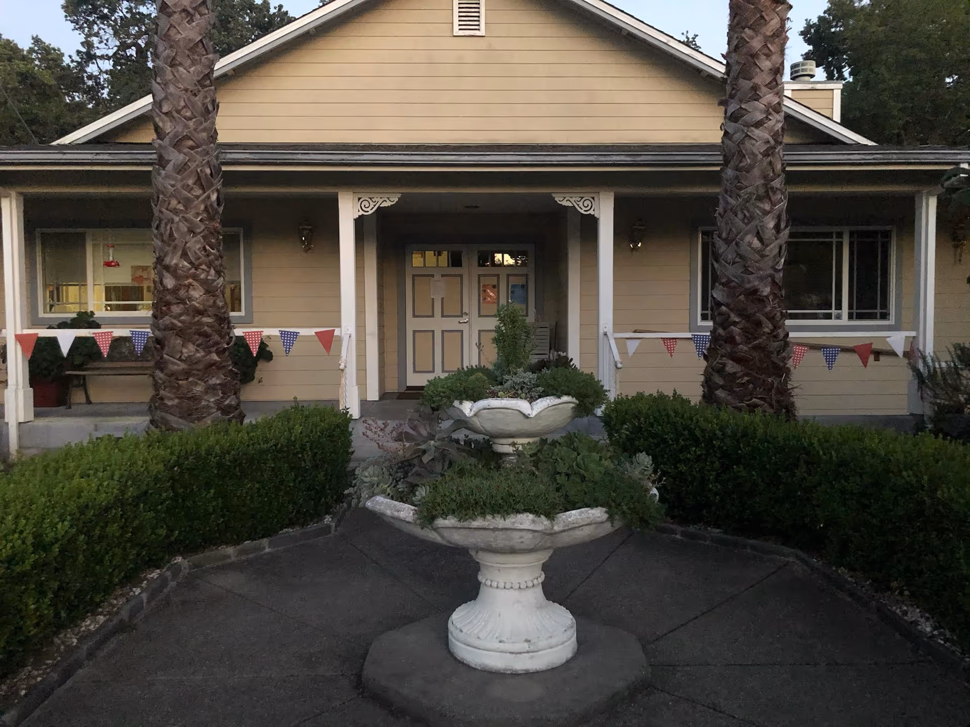 Front entrance of a single-story building with a central tiered fountain, two tall palm trees, and decorative bunting.