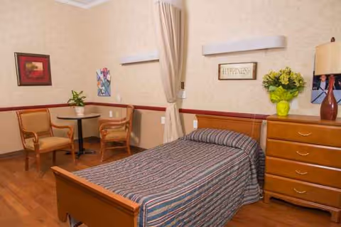 Cozy bedroom in a senior living facility with bed, dresser, curtains, and seating.
