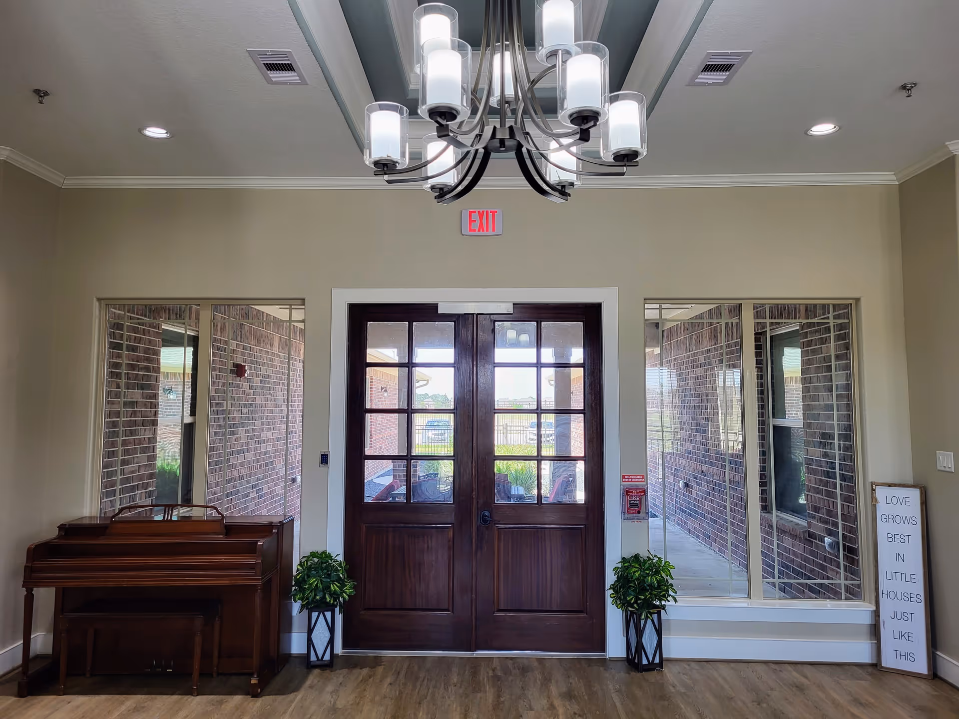 Interior view of a room with double wooden doors featuring glass panels. On either side of the doors are large windows showing a brick exterior wall. A wooden piano with a bench is placed to the left, and two potted plants are positioned on either side of the doors. A chandelier with multiple lights hangs from the ceiling. A sign on the right reads 'LOVE GROWS BEST IN LITTLE HOUSES JUST LIKE THIS'.