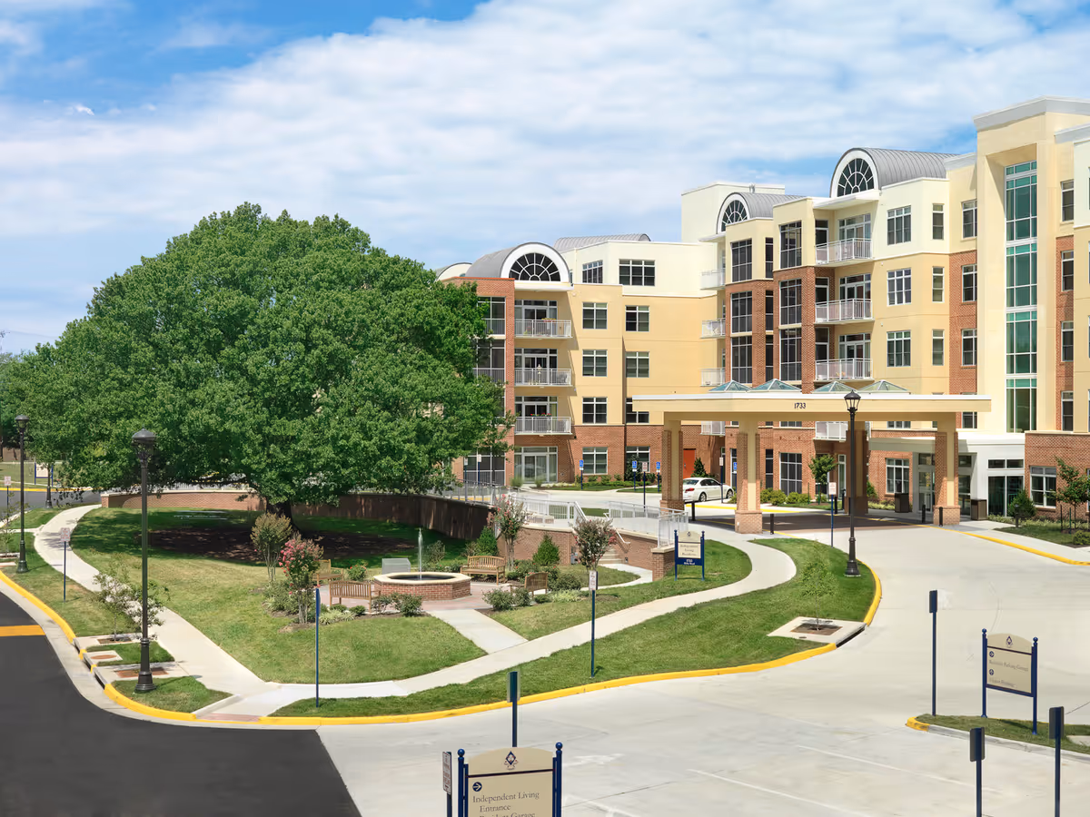 Exterior view of The Sylvestery at Vinson Hall Retirement Community showing a multi-story building with a covered entrance, large windows, and balconies. In front of the building is a landscaped area with a large tree, benches, a circular brick planter, and paved walkways. The sky is partly cloudy.