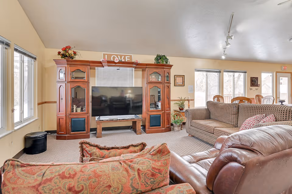 A cozy living room in an assisted living facility featuring a large wooden entertainment center with a flat-screen TV, a patterned sofa, a leather recliner, and a cushioned armchair. The room has beige walls, carpeted floor, several windows letting in natural light, and decorative plants and artwork.