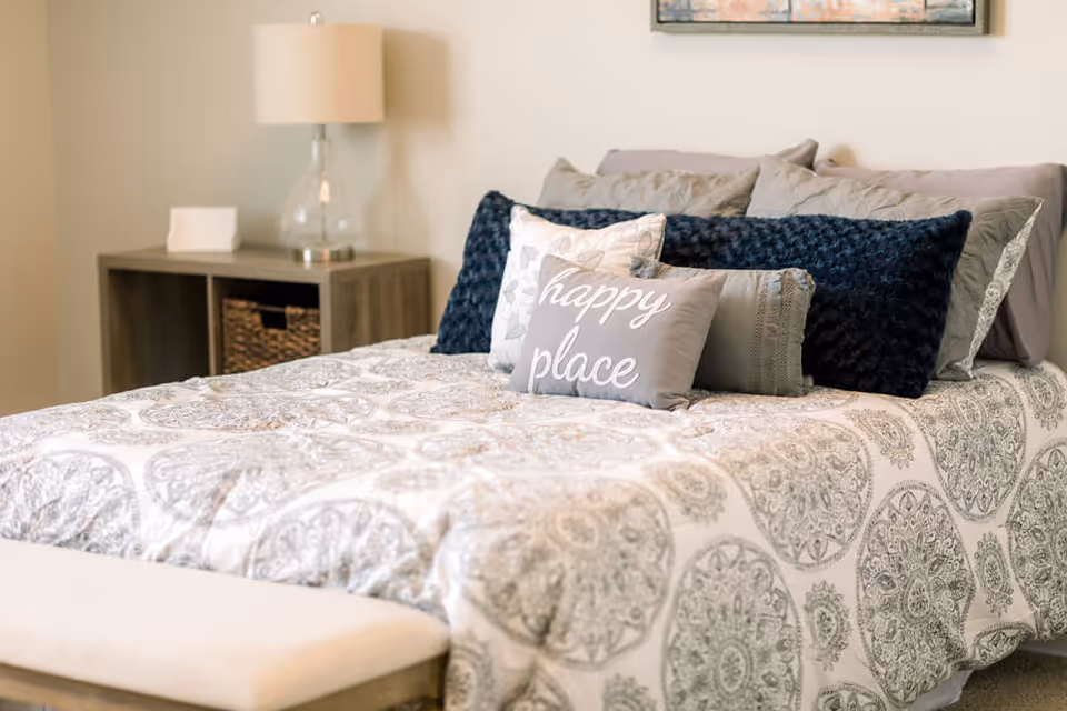 A neatly made bed with patterned bedding and multiple pillows, including one with the words 'happy place'. A wooden nightstand with a lamp and a small decorative item is beside the bed. The room has a calm and cozy atmosphere.