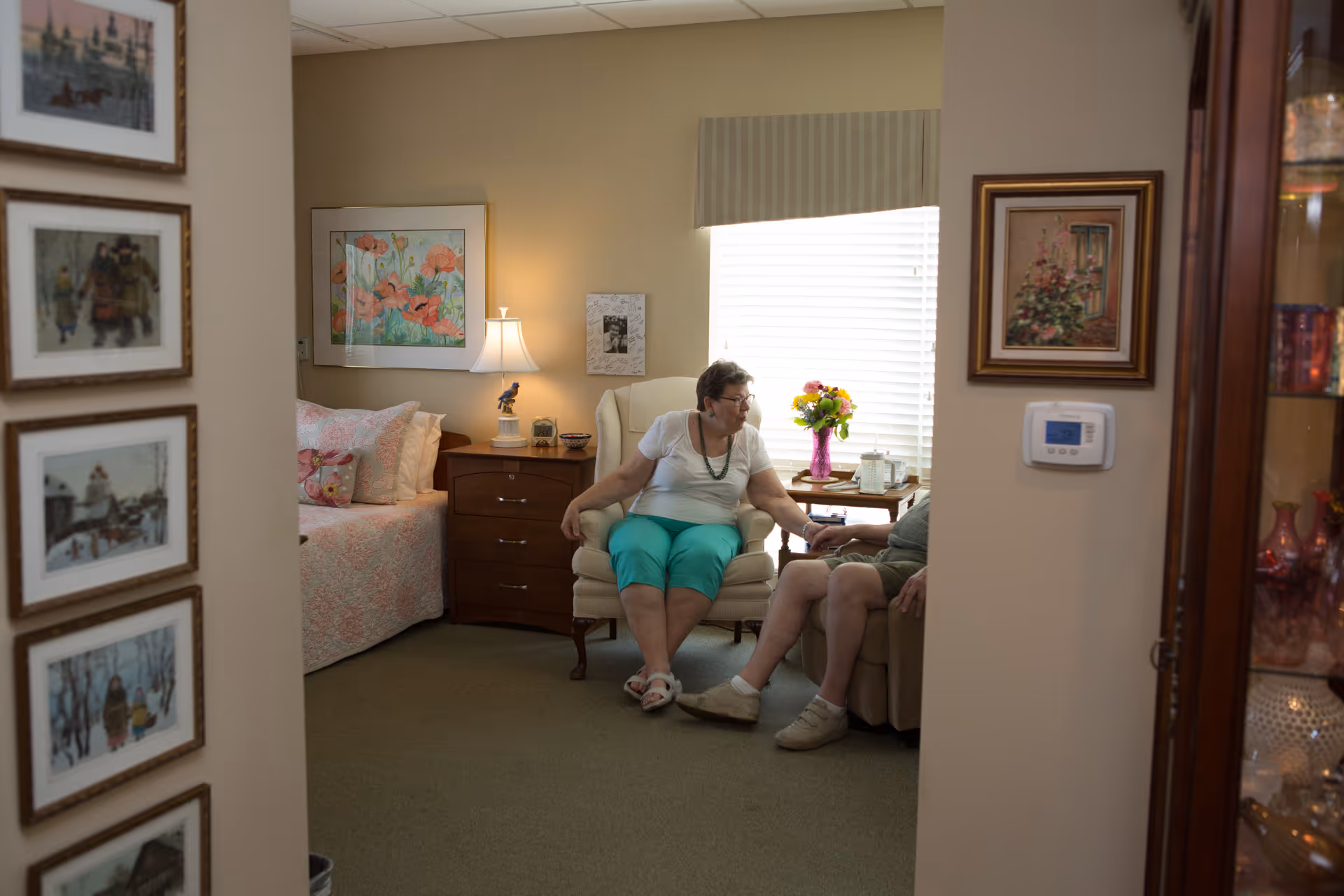 Two elderly people sitting and holding hands in a cozy bedroom with a bed, nightstand, lamp, and framed artwork on the walls. A window with blinds and a vase of flowers on a side table is visible in the background.