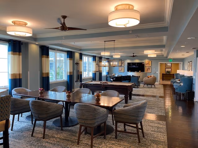 Bright communal senior living room with dining tables and chairs in the foreground, a pool table and lounge seating toward the back, and large windows along one wall.