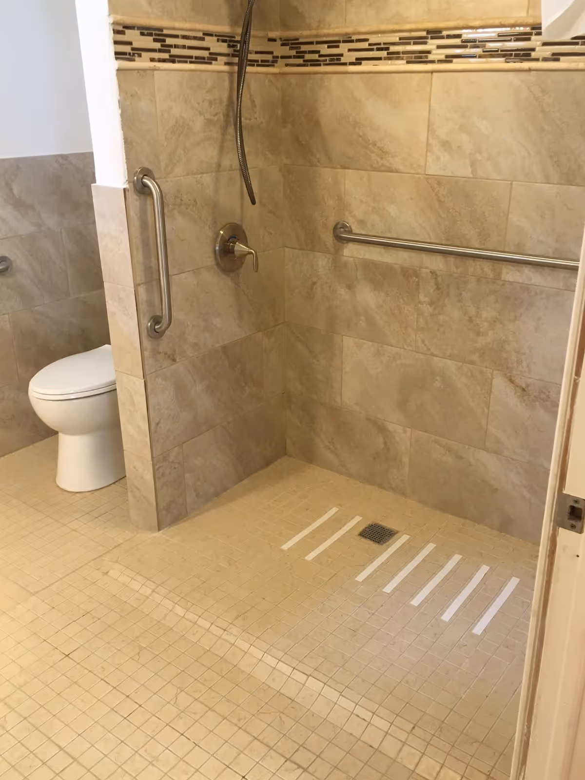 A bathroom with beige tiled walls and floor, featuring a walk-in shower area with a handheld showerhead, grab bars on the wall, and white anti-slip strips on the shower floor. A white toilet is visible in the background.