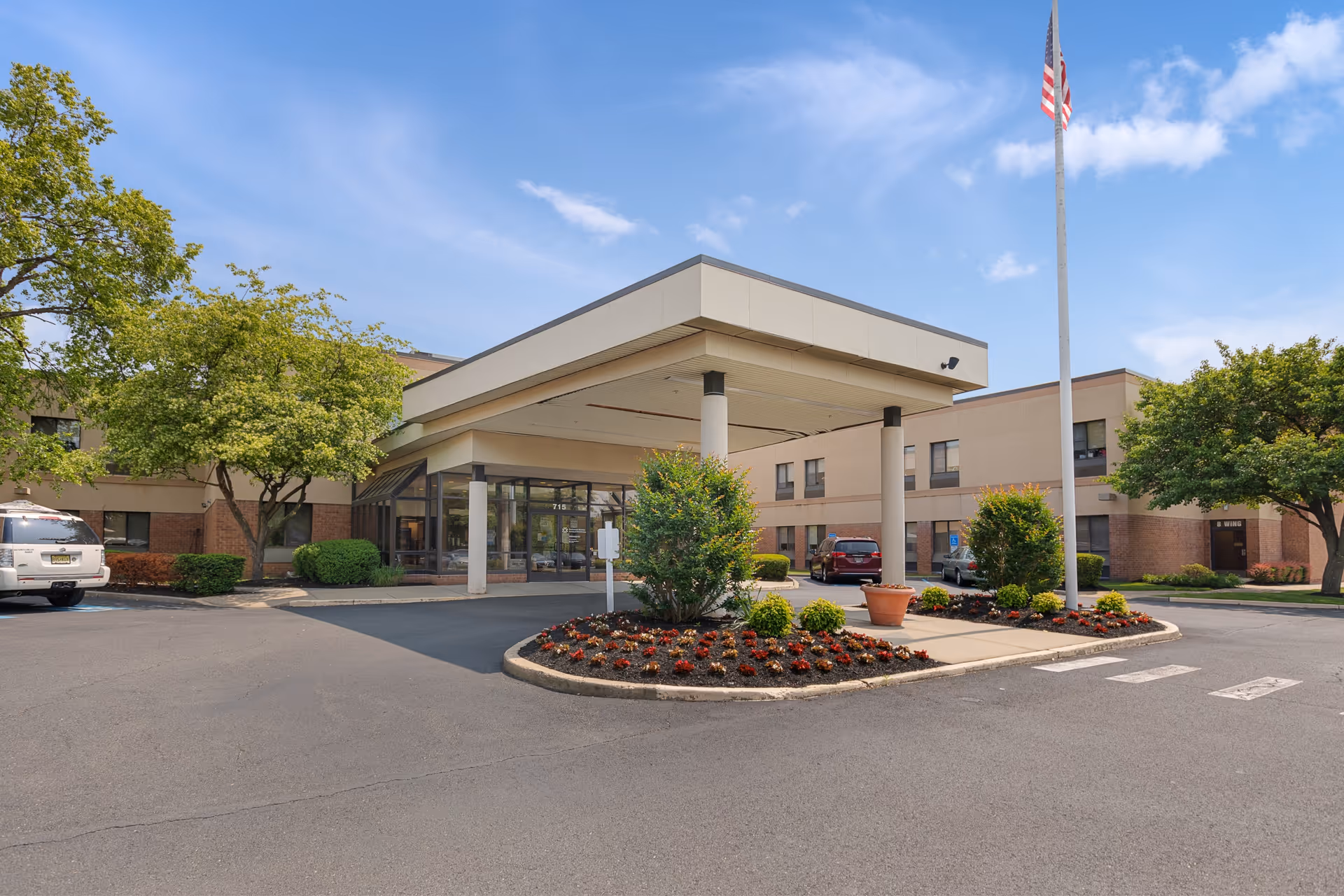 Exterior view of a senior living facility entrance with a covered drop-off area, landscaped flower beds, trees, parked cars, and an American flag on a flagpole under a blue sky.