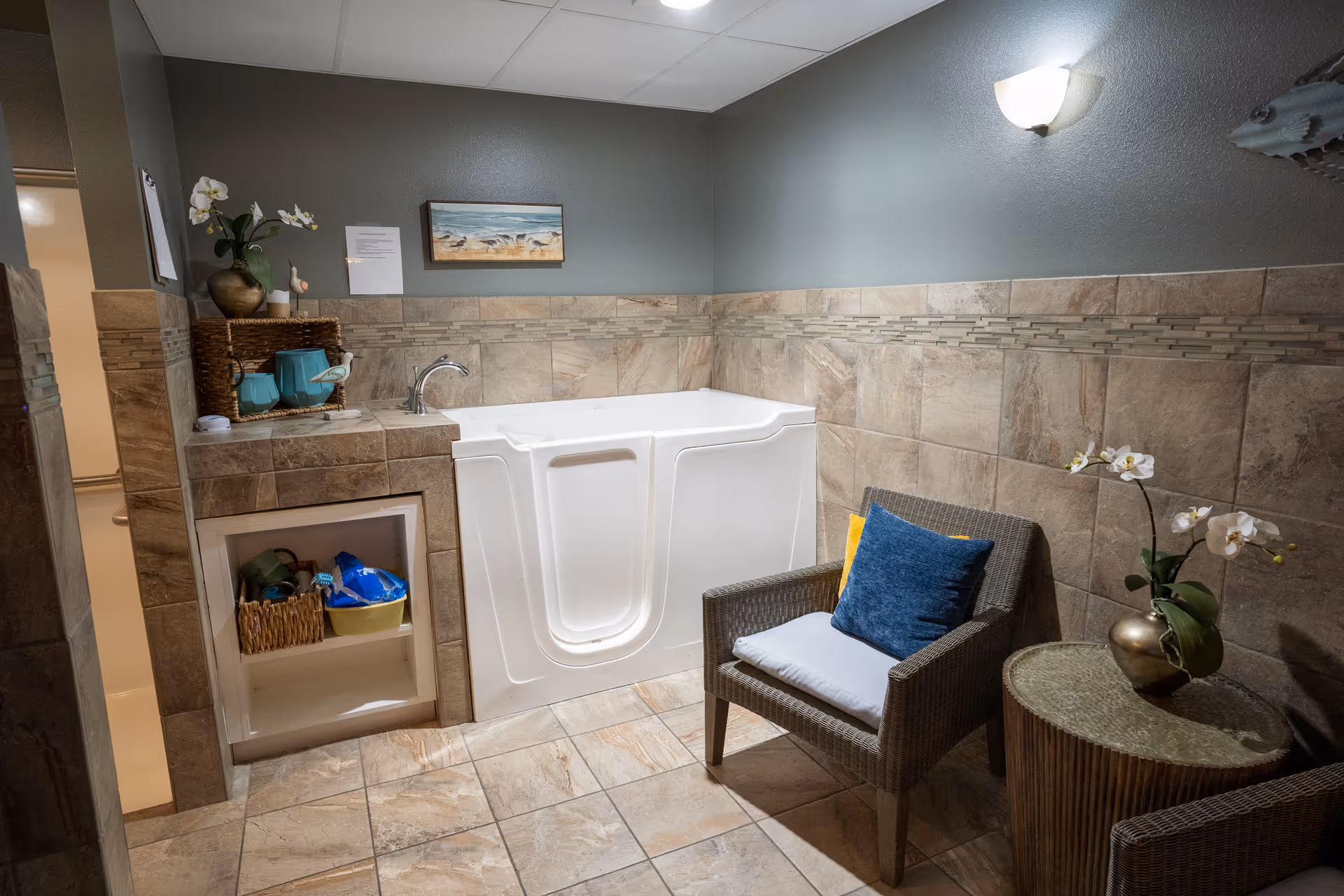A tiled bathroom with a walk-in bathtub, a wicker chair with cushions, a small round table with a potted orchid, and decorative items on a shelf. The walls are tiled halfway up and painted gray above, with a wall-mounted light fixture and a small framed beach painting.