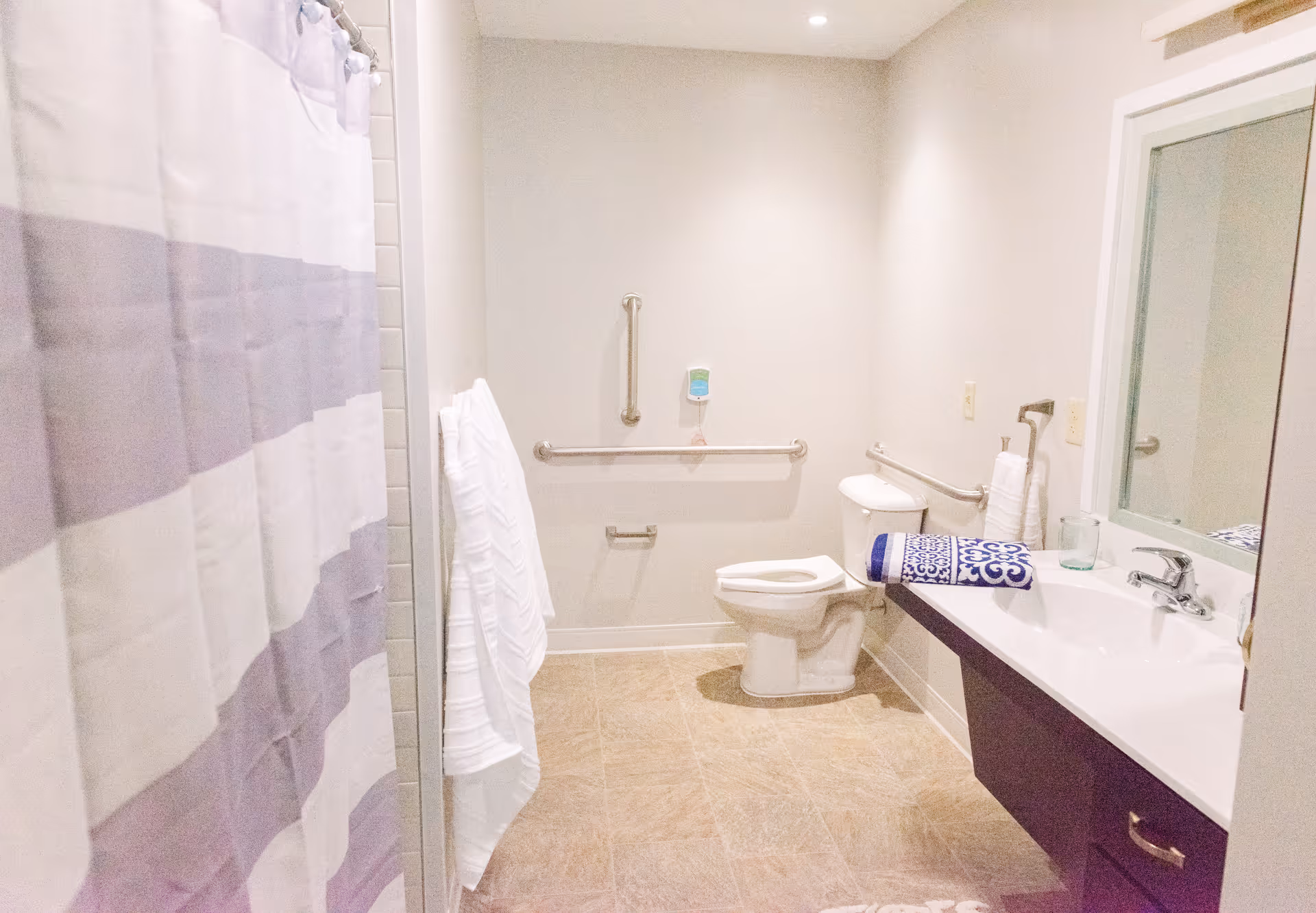 A clean and accessible bathroom with a toilet, a sink with a large mirror, grab bars on the walls, a shower with a striped shower curtain, and neatly folded towels.