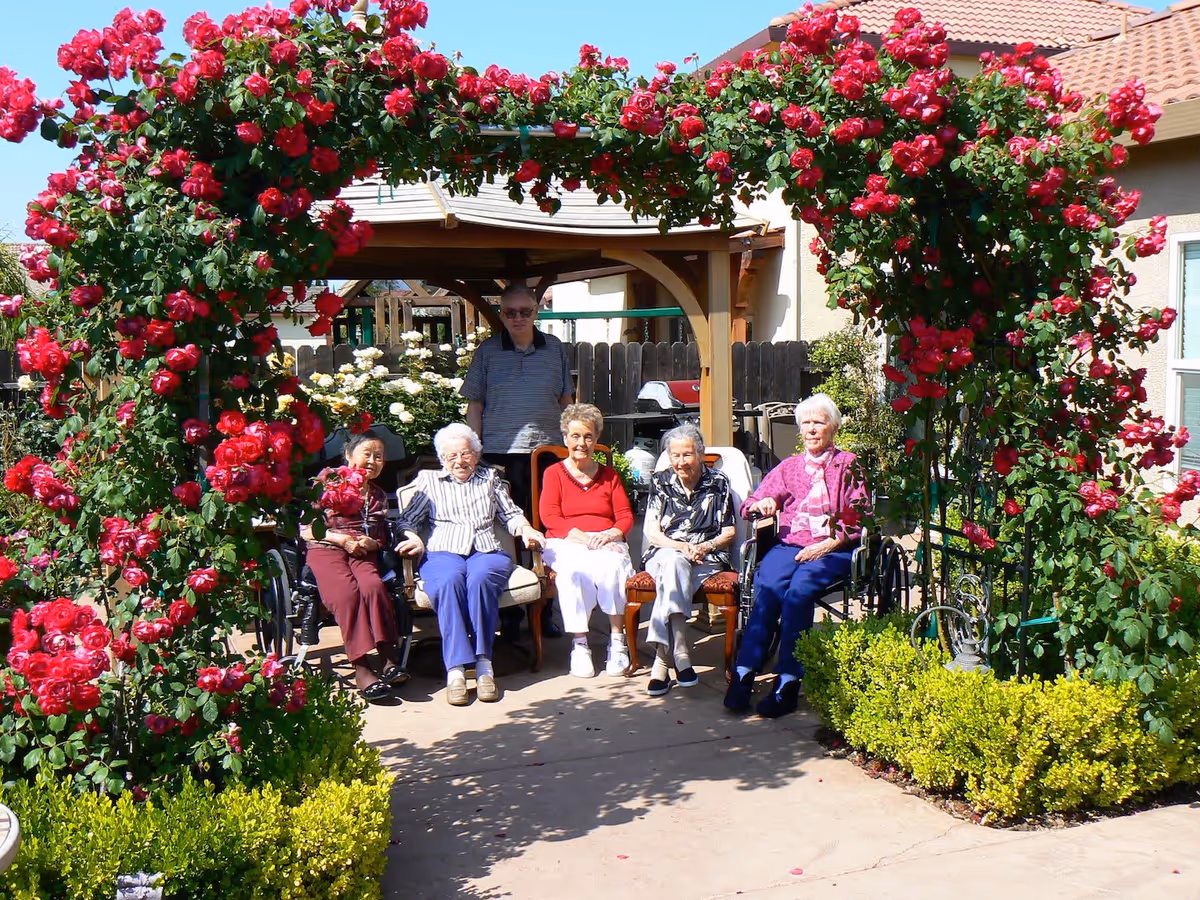 A group of elderly people sitting and standing under a wooden pergola in a garden area with vibrant red roses climbing over an arch. The setting is sunny with clear blue skies, and there are green bushes and a building with a tiled roof in the background.
