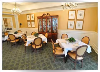 Dining room with white-tablecloth tables, chairs, a china cabinet, and framed artwork.