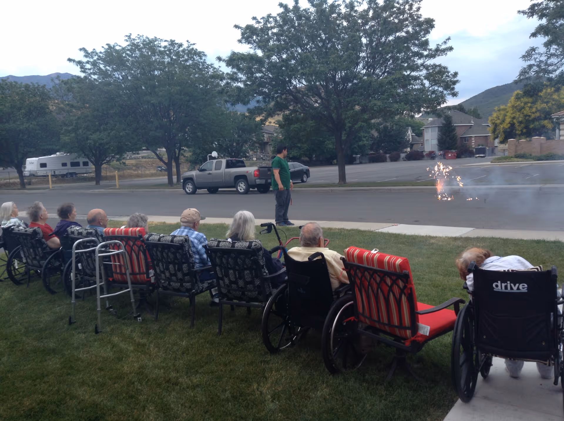 A row of people in wheelchairs and chairs on a lawn watching small fireworks in the street as a person stands nearby.