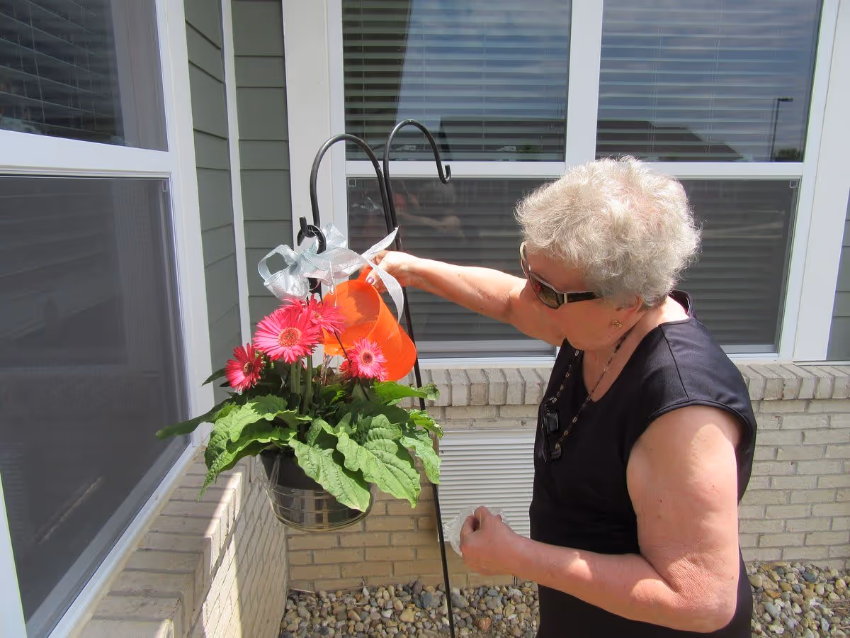 An elderly woman wearing sunglasses and a sleeveless black top is watering a hanging pot of pink flowers outside a building with beige brick walls and large windows.