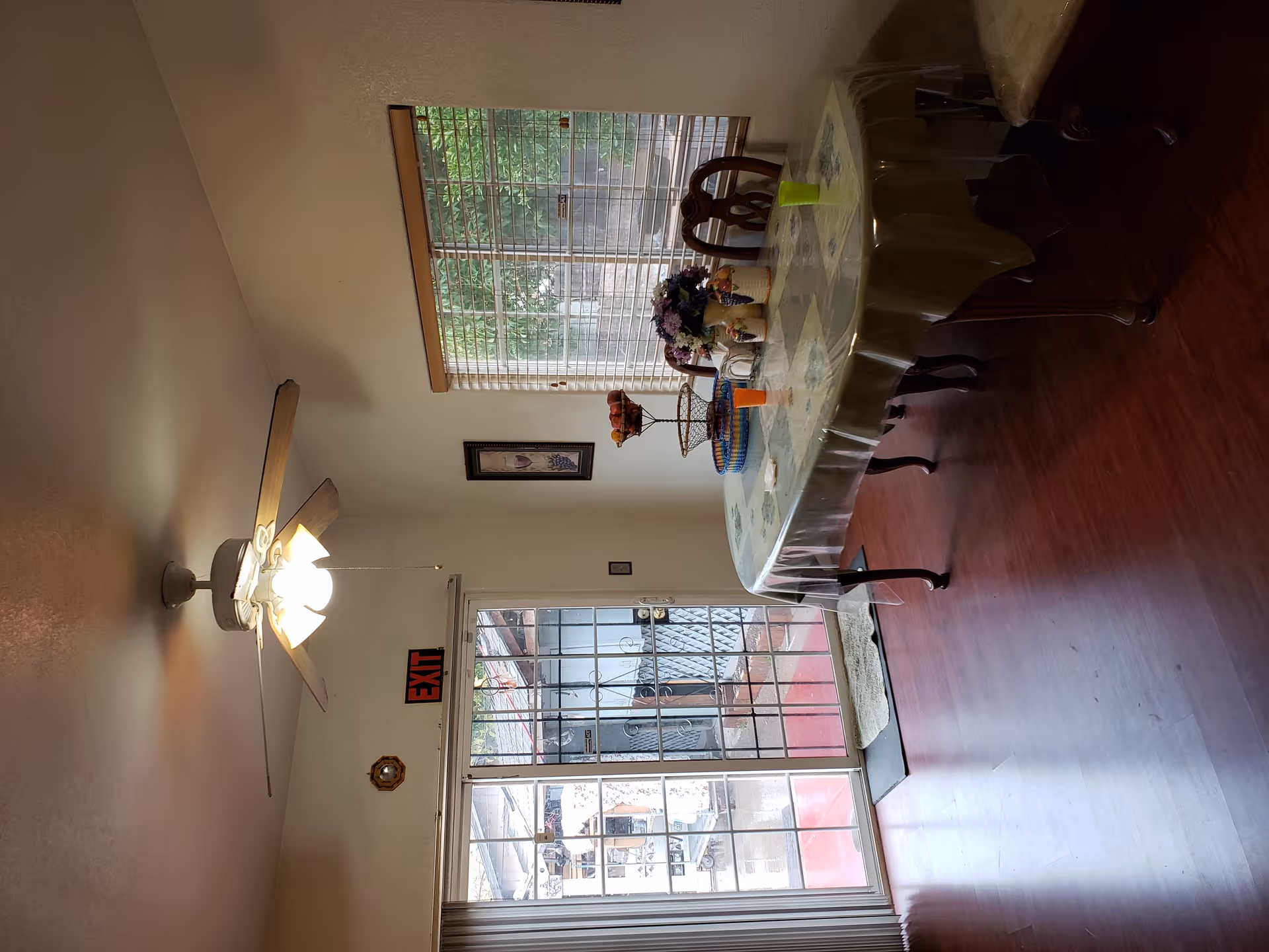 Interior view of a dining area with a table covered by a plastic tablecloth, surrounded by chairs. There is a ceiling fan with lights turned on, a window with blinds, and a glass door leading outside. The floor is wooden, and there is an exit sign above the door.