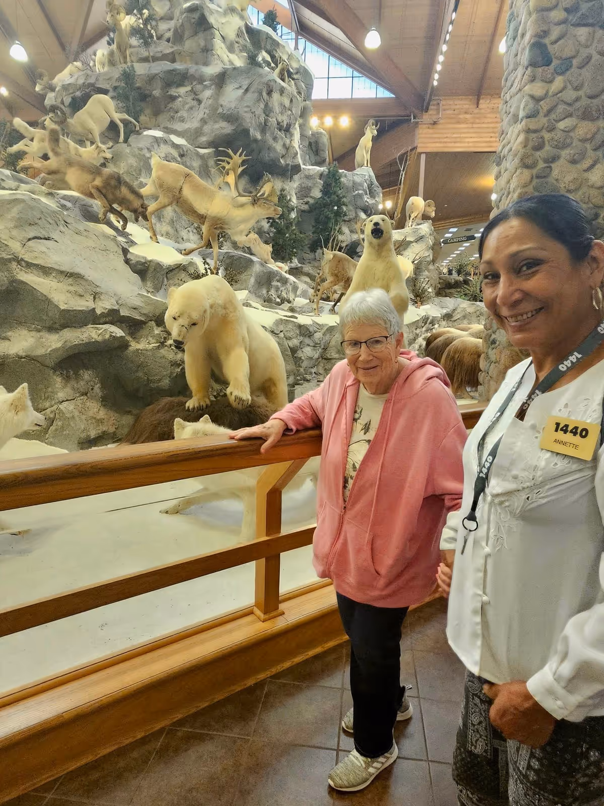 An elderly woman in a pink hoodie and glasses stands next to a smiling woman wearing a white blouse and a name tag that reads '1440 Annette.' They are indoors near a wooden railing, with a large display of taxidermy animals including polar bears, deer, and wolves on rocky terrain behind them.