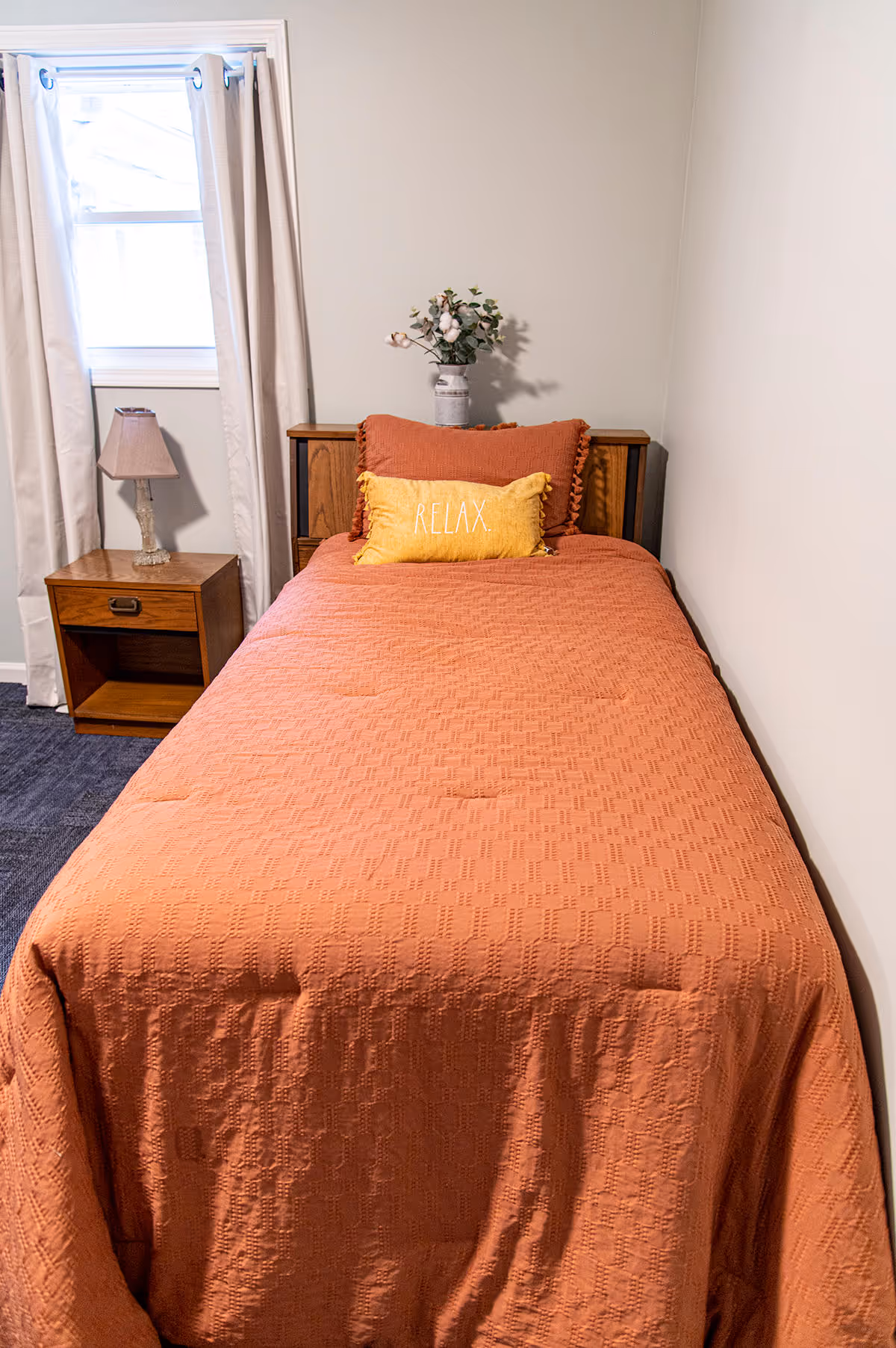 A small bedroom with a single bed covered in an orange textured bedspread. There are two pillows on the bed, one orange and one yellow with the word 'RELAX' embroidered on it. A wooden nightstand with a lamp is positioned next to the bed, and a window with beige curtains is visible behind the nightstand. A small vase with flowers sits on the headboard.