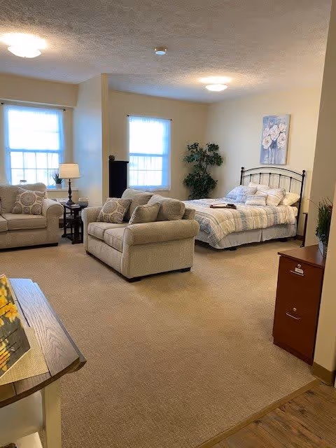 A cozy living space featuring a bed with striped bedding, two beige upholstered armchairs, a matching loveseat, a side table with a lamp, two windows with white curtains, a potted plant, and a floral painting on the wall.