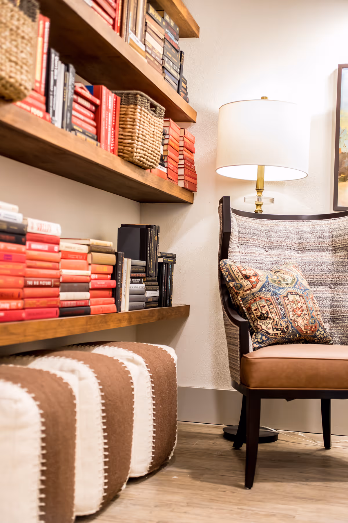 Cozy reading nook with wooden shelves filled with books and woven baskets, a cushioned armchair with a patterned pillow, a standing lamp with a white shade, and two brown and white striped poufs on a wooden floor.