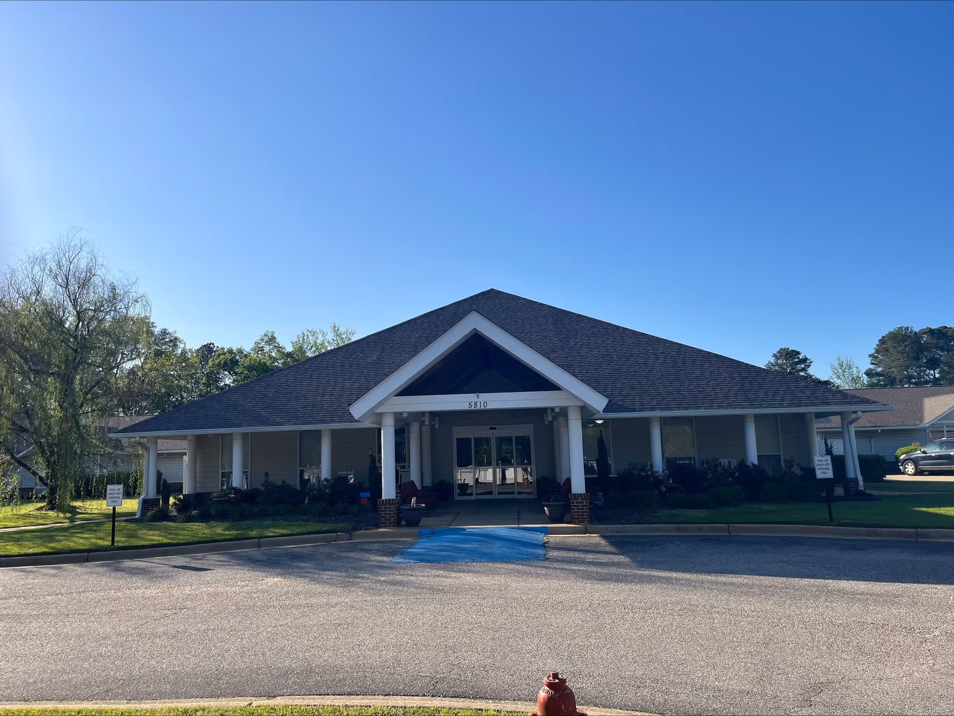 Front exterior view of a single-story building with a dark shingled roof and white columns supporting a covered entrance. The building is surrounded by green grass, bushes, and trees under a clear blue sky. There is a blue handicapped parking space and a red fire hydrant in front of the building.