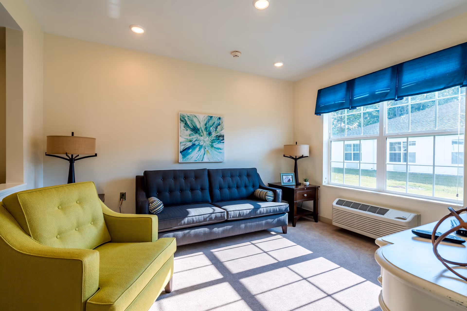 A bright living room with a large window covered by a blue valance, letting in sunlight that casts shadows on the carpeted floor. The room features a dark blue tufted sofa with two striped cylindrical pillows, a green upholstered armchair, two matching table lamps on dark wooden side tables, a framed photo, a small plant, and a piece of abstract artwork with blue and green tones hanging on the wall.