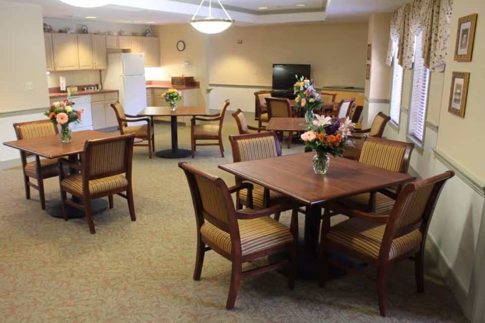 A dining area in an assisted living facility with several wooden tables, each surrounded by four striped cushioned chairs. Each table has a vase with colorful flowers. In the background, there is a kitchenette with cabinets, a refrigerator, and a dishwasher. A television is visible on a stand against the far wall. The room has carpeted flooring and windows with patterned valances.