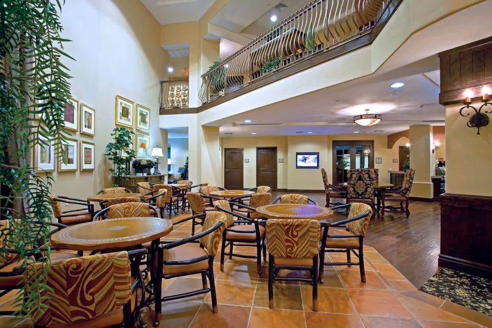 Two-story interior dining/lounge area with multiple round tables and upholstered chairs beneath a balcony and warm lighting.