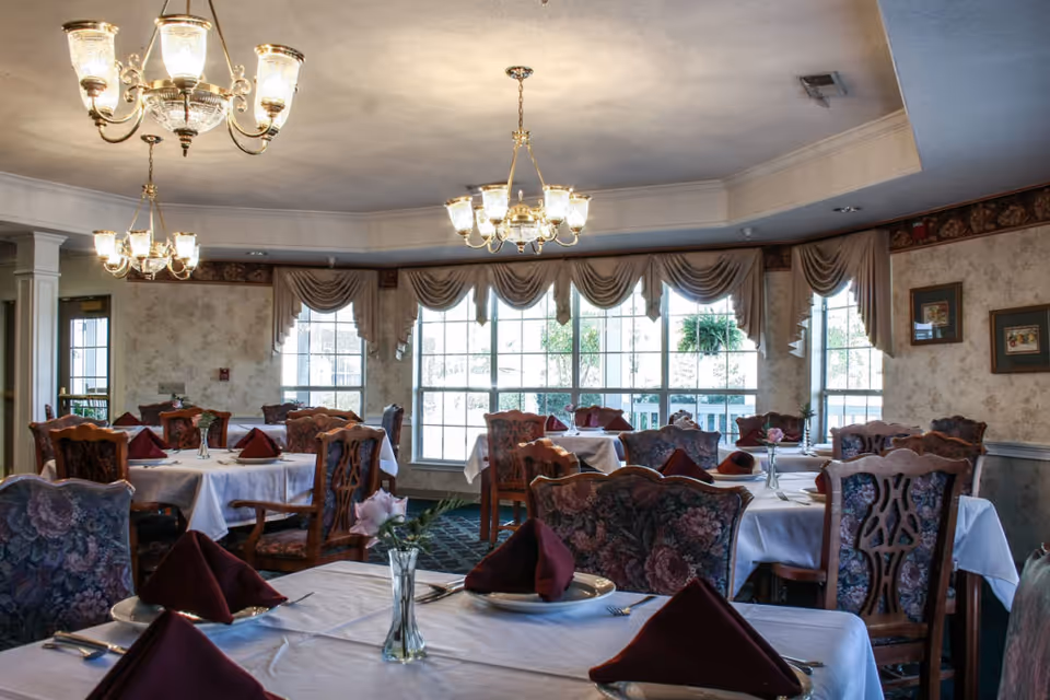 Dining room with neatly set tables, chandeliers, floral-upholstered chairs, and large windows.