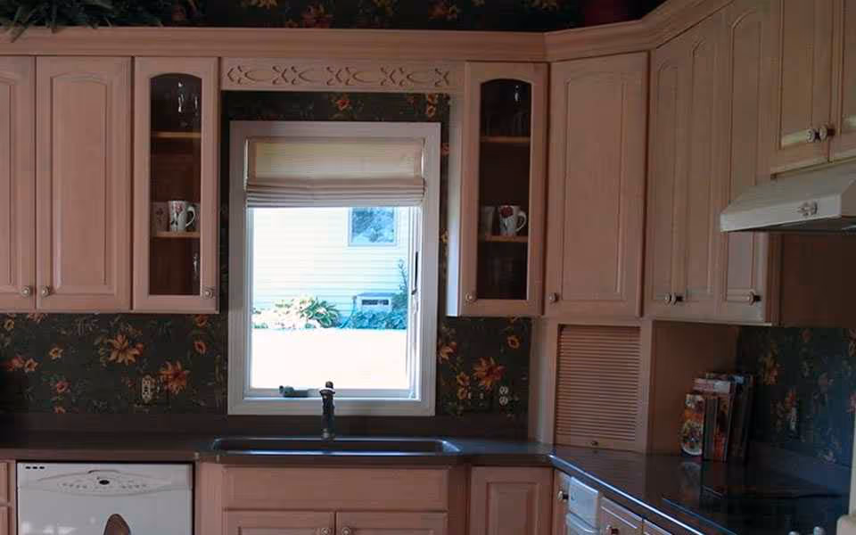 A kitchen interior featuring light pink cabinets with glass doors on the upper cabinets, a window with a white blind above a sink, floral wallpaper, a dishwasher, and a stovetop with a range hood.