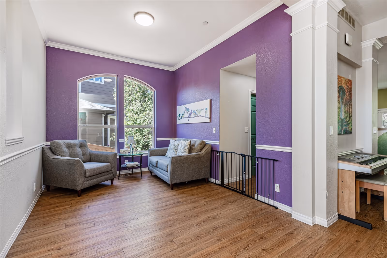 A cozy sitting area with two gray upholstered chairs and a small round table between them, placed against a purple accent wall with a large window showing greenery outside. The room has wooden flooring and white trim, with a baby gate installed in the doorway leading to another room.