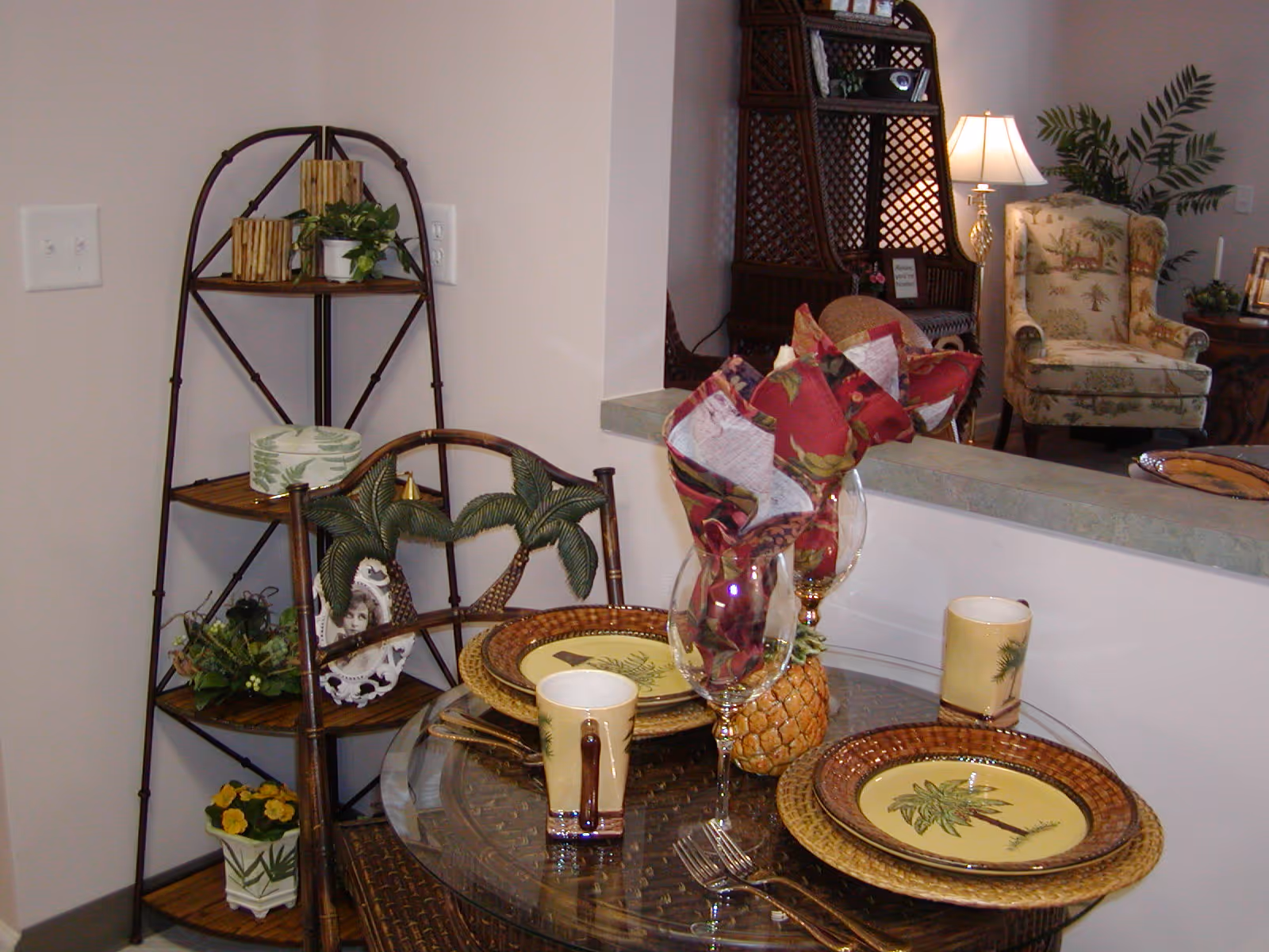 A small dining area with a round glass table set for two, featuring palm tree-themed plates and mugs, wine glasses with red and white patterned napkins, and a decorative pineapple centerpiece. Behind the table is a corner shelf with plants and decorative items. In the background, there is a cozy seating area with a patterned armchair, a lamp, and a plant.