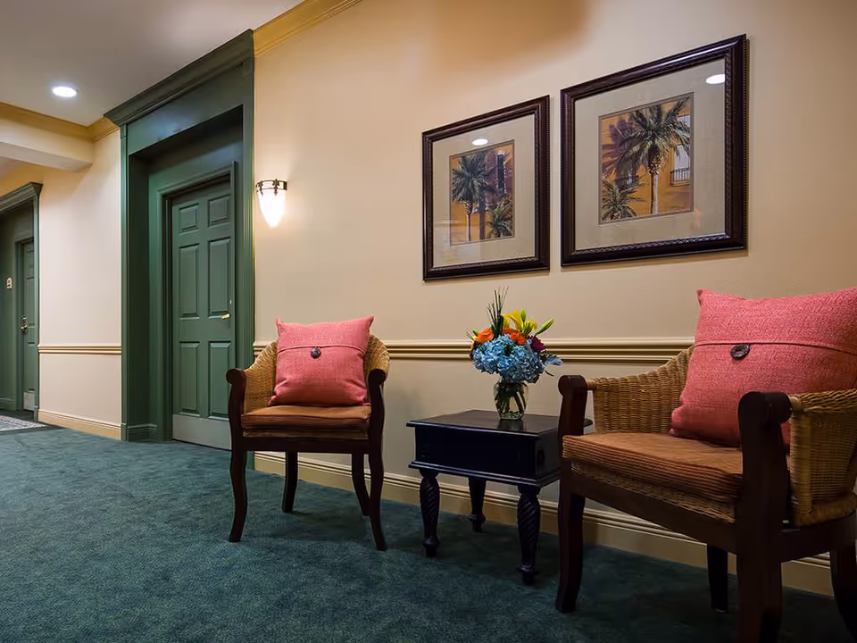 A carpeted interior hallway seating area with two chairs topped by pink pillows, a small side table with a vase of flowers, framed artwork, and green doors.