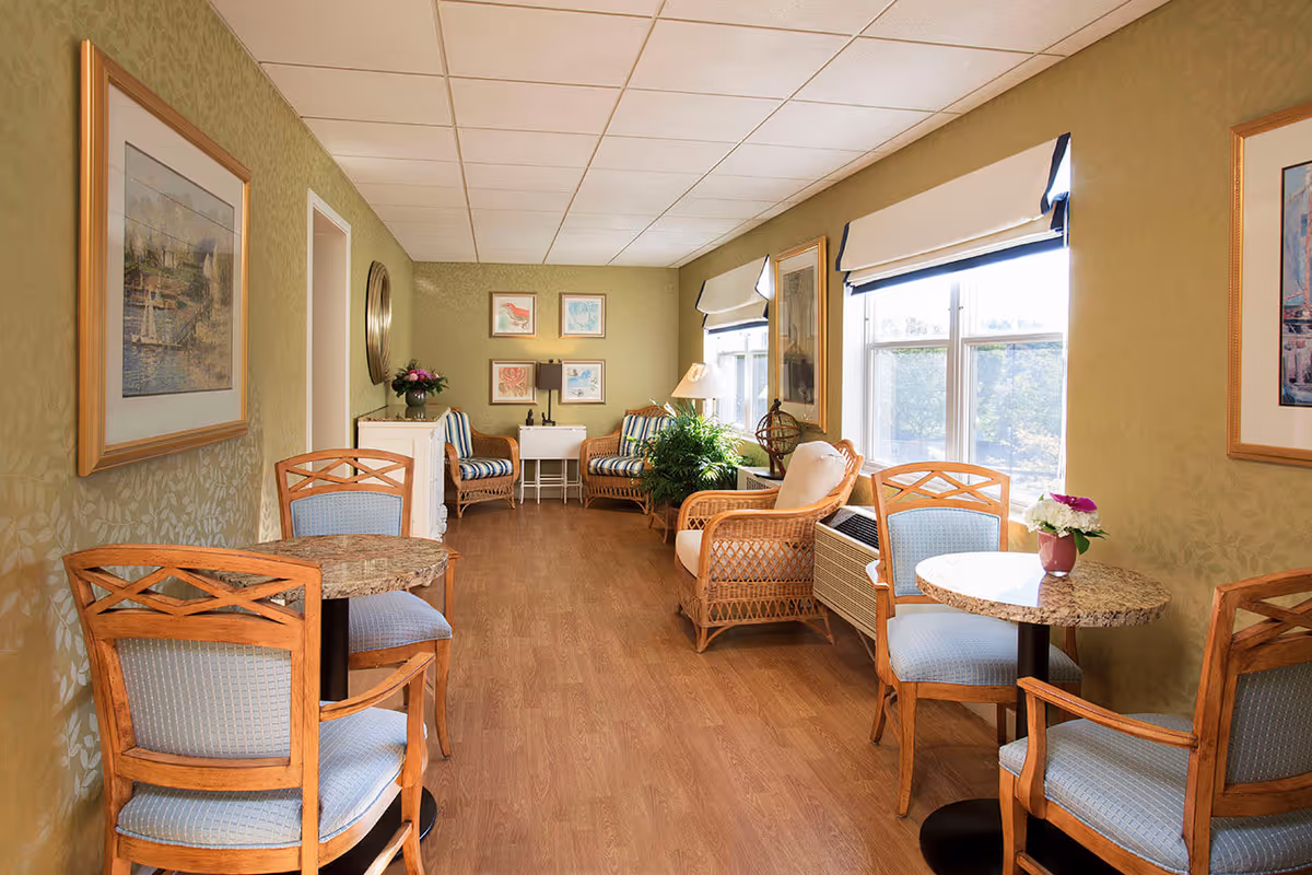 A cozy sitting area in a senior living facility with wooden chairs upholstered in light blue fabric around small round tables. The room has light green patterned walls, large windows with white and blue valances, and wicker armchairs with cushions. The floor is wood, and the walls are decorated with framed artwork and a round mirror. There are also plants and a lamp adding to the inviting atmosphere.