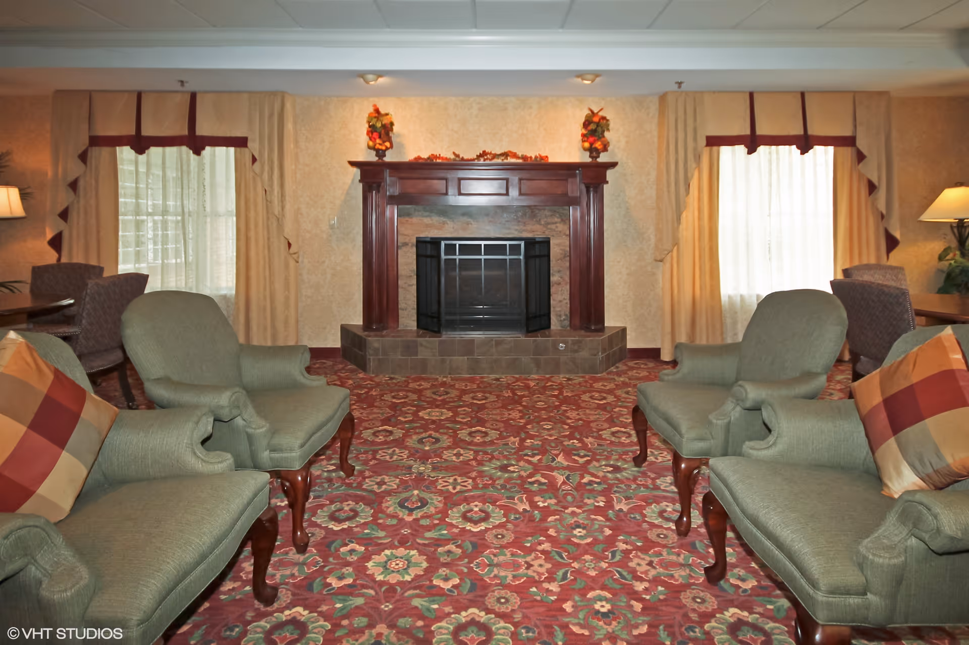A cozy living room with a patterned red carpet, two green upholstered armchairs and two matching sofas with plaid pillows facing a wooden fireplace mantel decorated with autumn-themed floral arrangements. The room has two windows with beige curtains and valances, and lamps on side tables providing warm lighting.