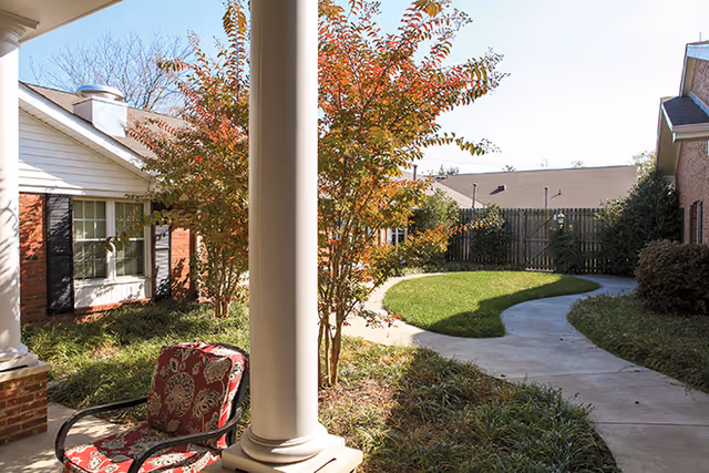 Covered courtyard with a column and patterned patio chair overlooking a winding paved walkway and landscaped lawn between brick buildings.