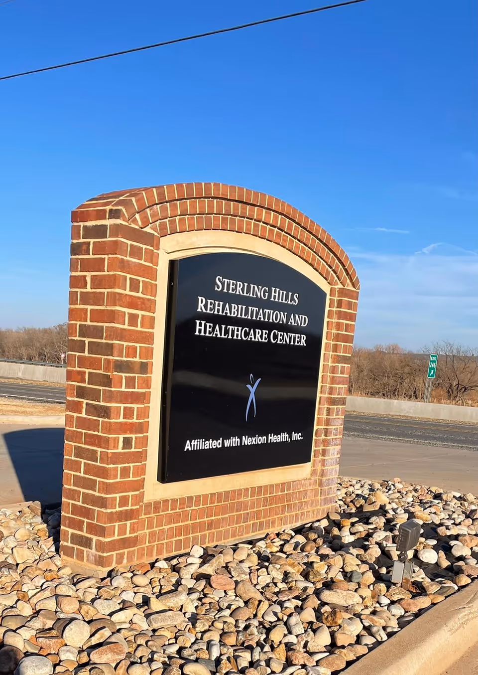Brick monument sign reading "Sterling Hills Rehabilitation and Healthcare Center" on a bed of rocks by the roadside under a blue sky.