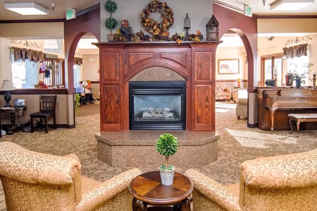 A cozy living room area with two patterned armchairs facing a wooden fireplace decorated with a wreath and small ornamental items. There is a small round wooden table with a potted plant between the chairs. The room has carpeted floors, arched doorways, and windows with valances letting in natural light. Additional seating and furniture are visible in the background.
