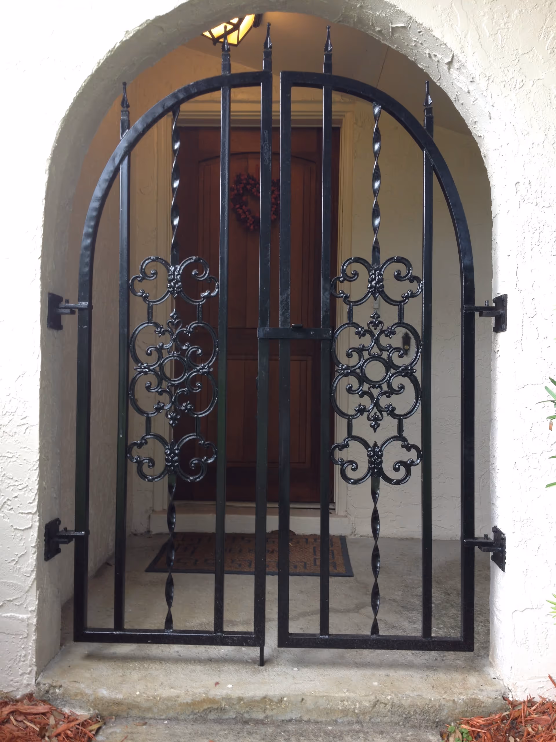 Black wrought iron gate with decorative scrollwork set within a white stucco archway, leading to a wooden front door with a wreath and a doormat on the concrete floor.