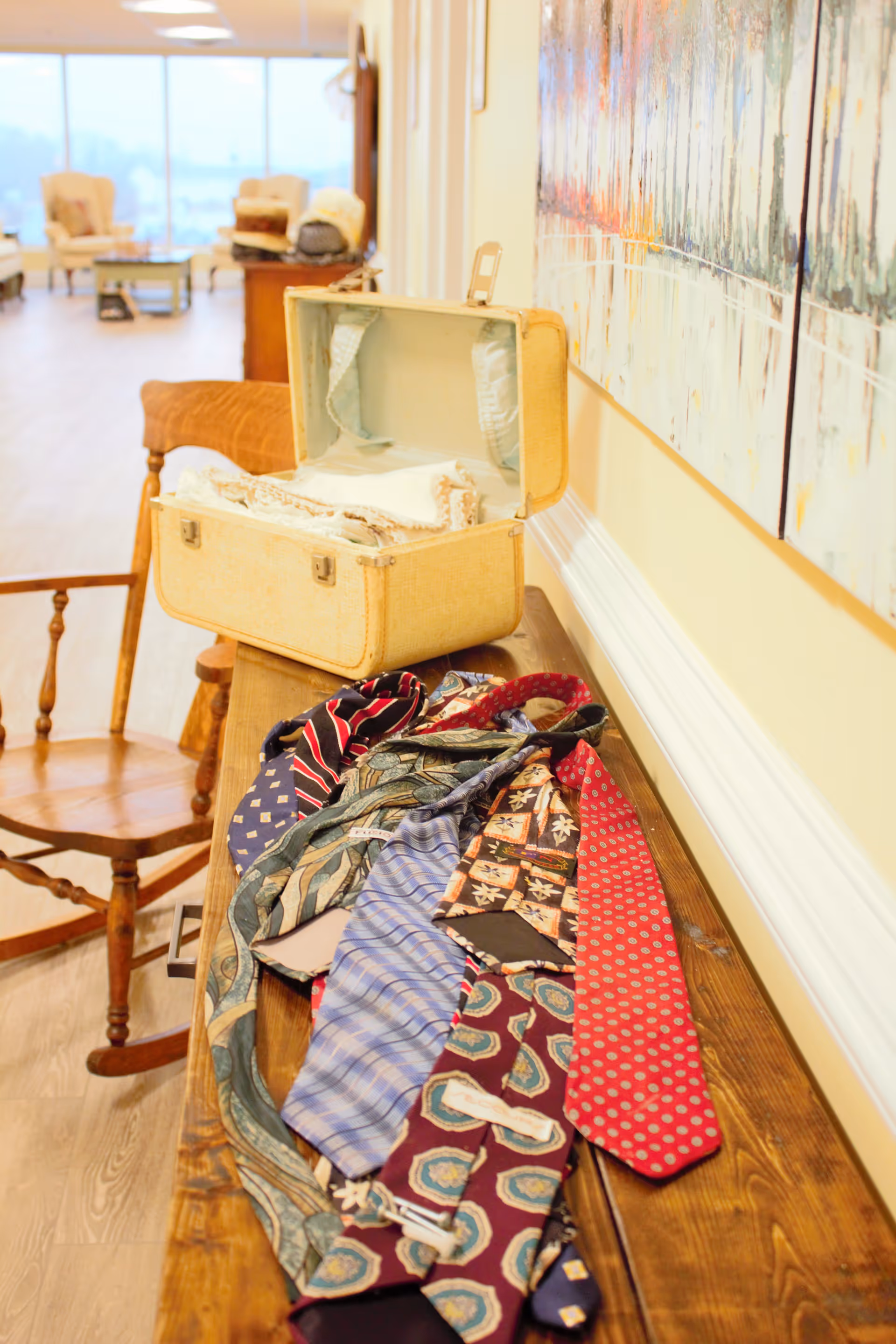 A wooden table with several colorful neckties laid out on it and an open vintage suitcase containing folded fabric. In the background, there is a wooden chair and a bright room with large windows and additional seating.