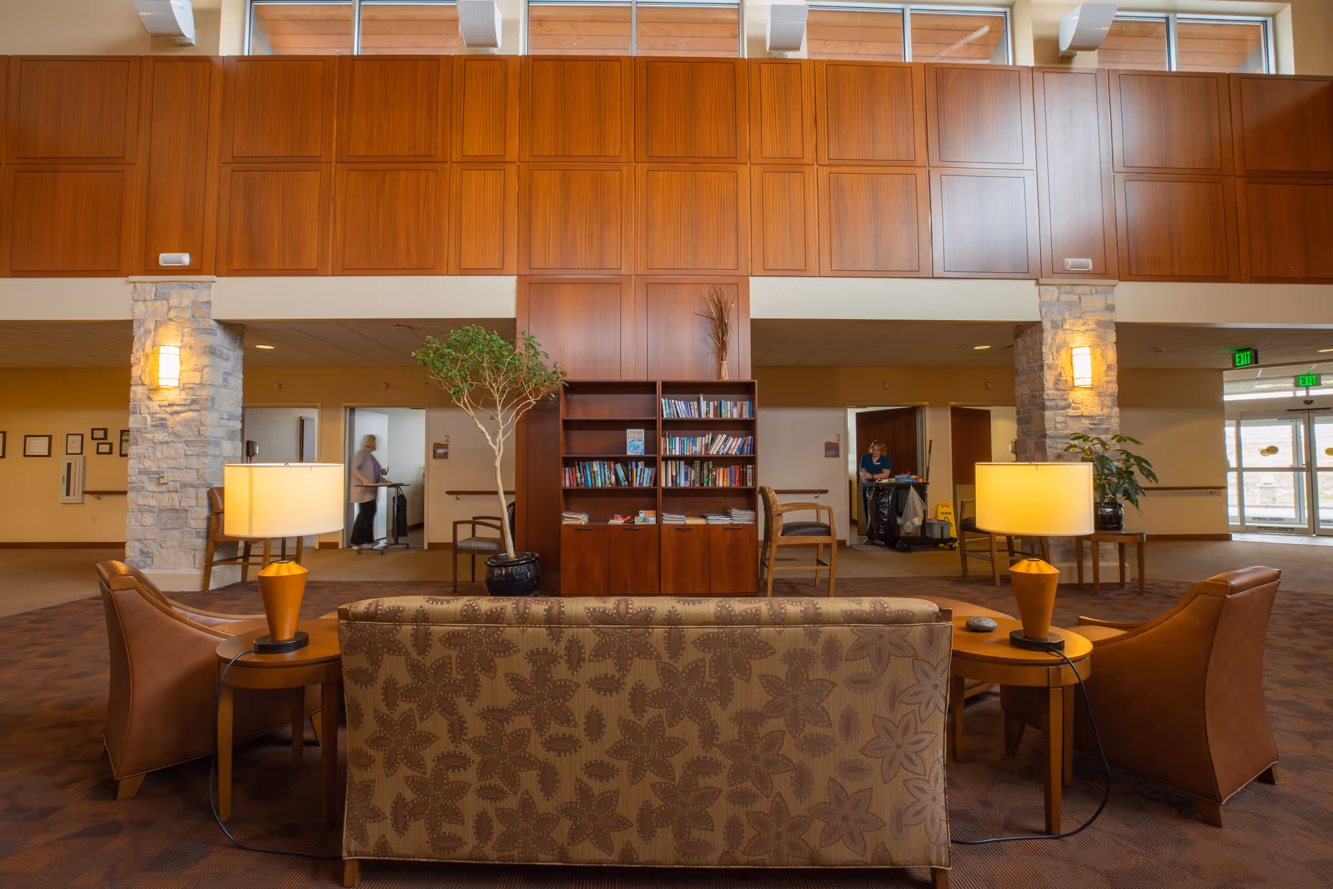 A spacious and well-lit common area in a senior living facility featuring a patterned sofa and two leather armchairs arranged around two wooden side tables with lamps. In the background, there is a wooden bookshelf filled with books, a potted plant, stone pillars with wall sconces, and two staff members visible in the hallway. Large windows near the ceiling allow natural light to enter the room.
